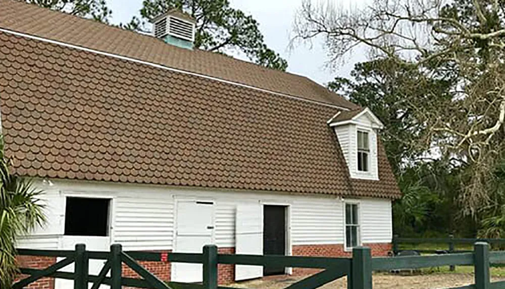 The image shows a white building with a brown shingle roof featuring a dormer window surrounded by a green fence with trees in the background