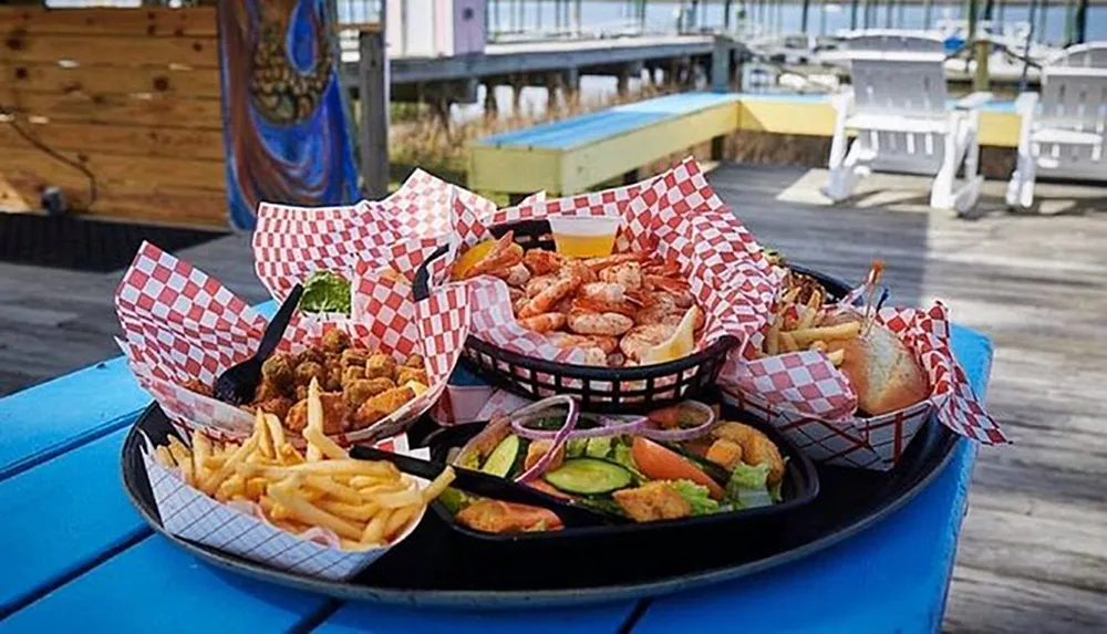 The image shows an array of seafood dishes served on a blue picnic table on a sunny waterfront deck