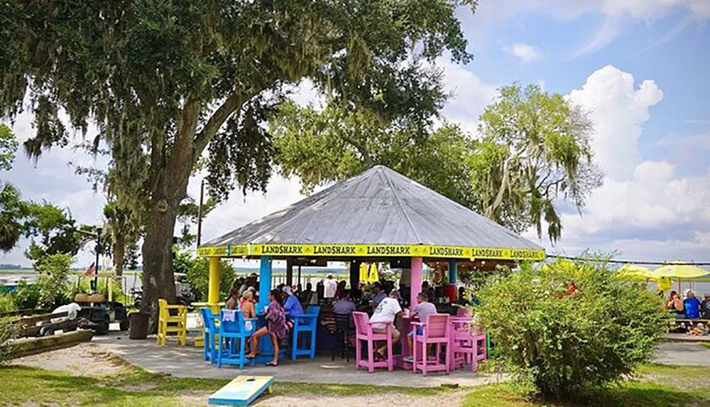 People are enjoying themselves at an outdoor bar with colorful furniture under the shade of trees and a gazebo-like structure with a view of the water and a blue sky peppered with clouds