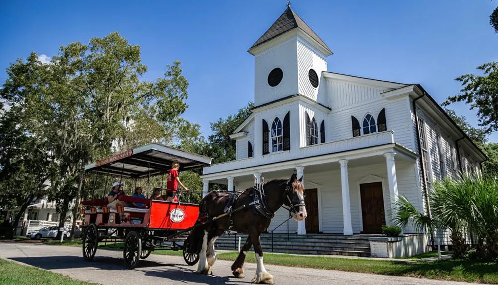 A horse-drawn carriage with passengers rolls by a white church building under a clear blue sky