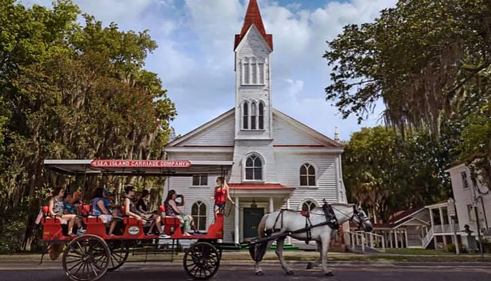 A horse-drawn carriage with passengers passes by an old white church with a tall steeple amidst lush greenery