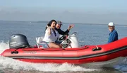 Three people are enjoying a ride on a red inflatable motorboat on a sunny day with clear skies.