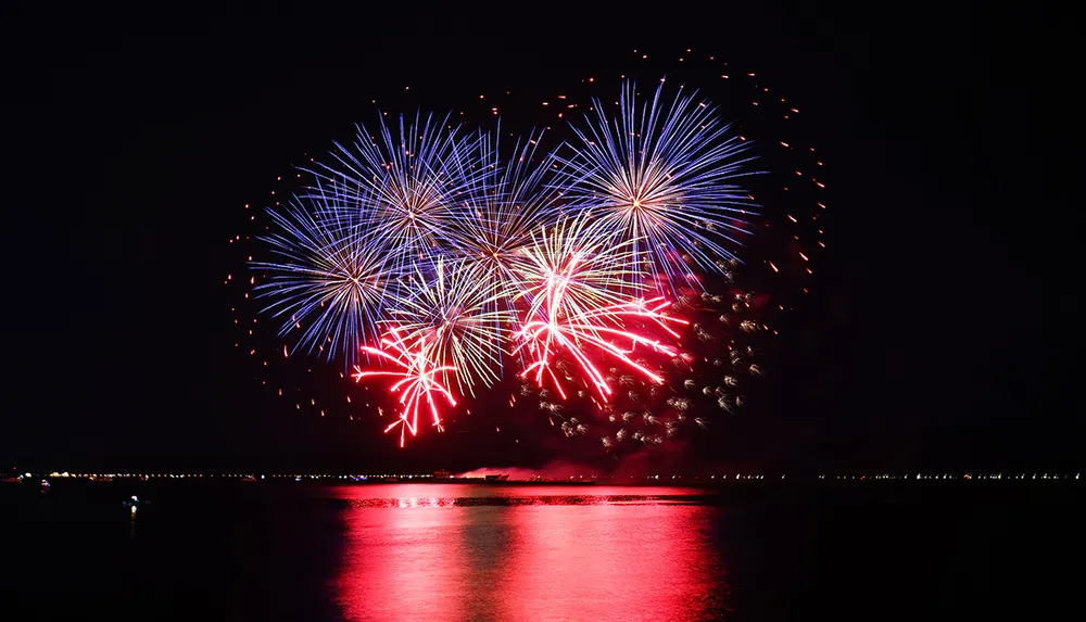 A vibrant display of red white and blue fireworks explodes over a reflective body of water at night
