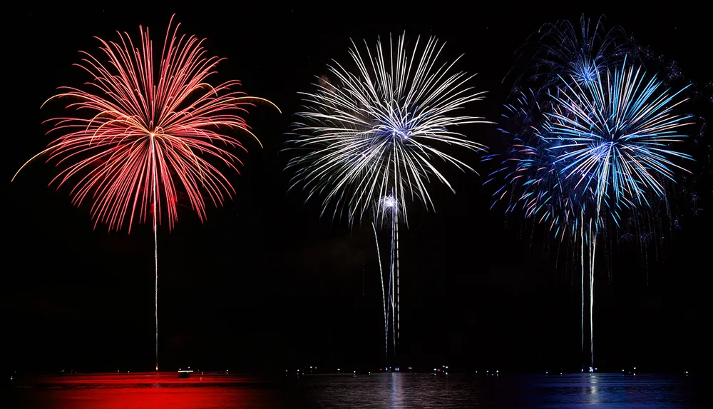 Three colorful fireworks are bursting in the night sky above a body of water reflecting on the surface below