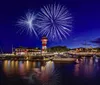 Vibrant fireworks illuminate the night sky above a quaint marina complete with a brightly colored lighthouse and docked boats