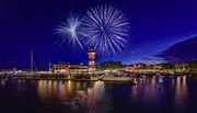 Vibrant fireworks illuminate the night sky above a quaint marina, complete with a brightly colored lighthouse and docked boats.