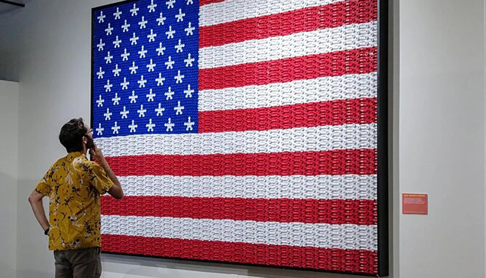 A person is observing a large American flag artwork made from what appears to be a collection of small items displayed on a gallery wall