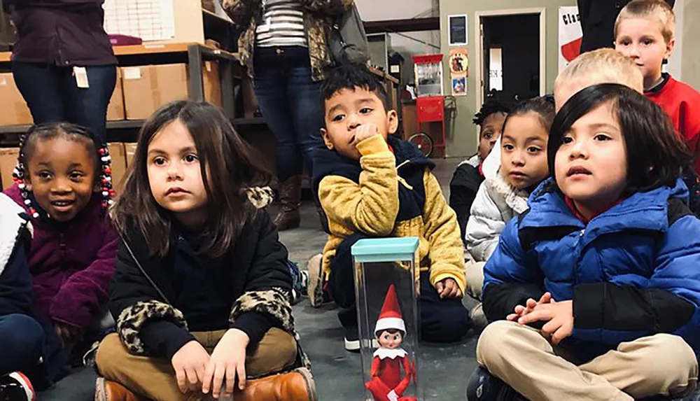A group of young children looks attentively at something out of frame with an Elf on the Shelf toy captured in the foreground
