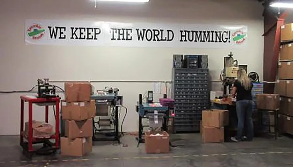 The image shows an industrial or warehouse setting with a banner that reads WE KEEP THE WORLD HUMMING and a person who appears to be working among various pieces of equipment and stacked boxes