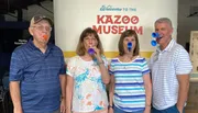 Four adults are playfully posing with colorful kazoos in their mouths in front of a sign that says 