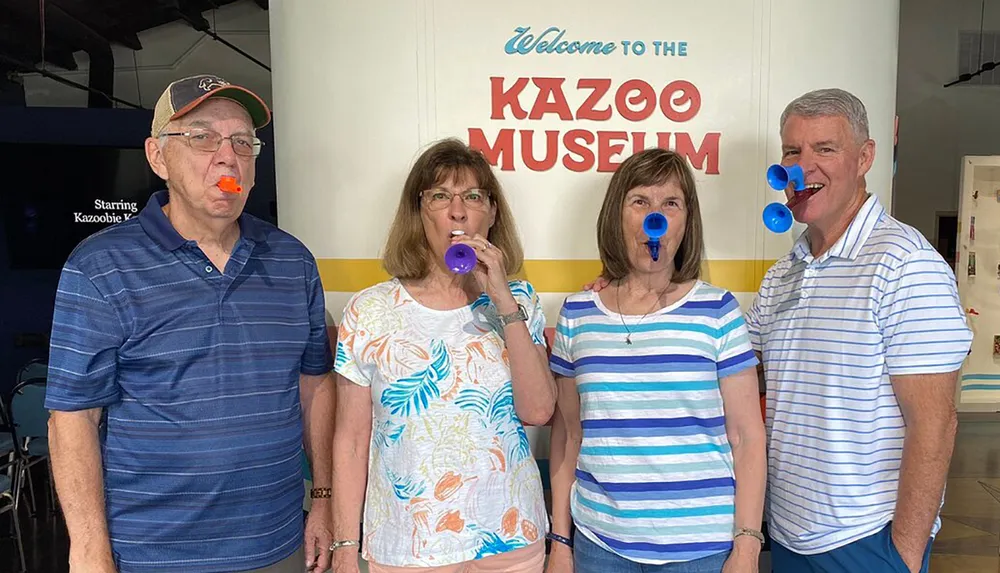 Four adults are playfully posing with colorful kazoos in their mouths in front of a sign that says Welcome to the Kazoo Museum