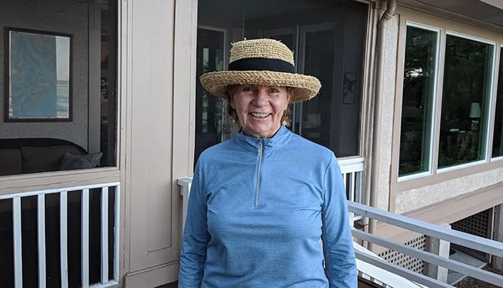 A smiling person wearing a straw hat and a blue zip-up top stands on a porch near a window and a white railing