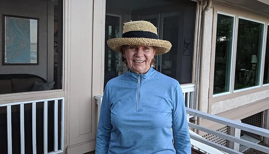 A smiling person wearing a straw hat and a blue zip-up top stands on a porch near a window and a white railing.