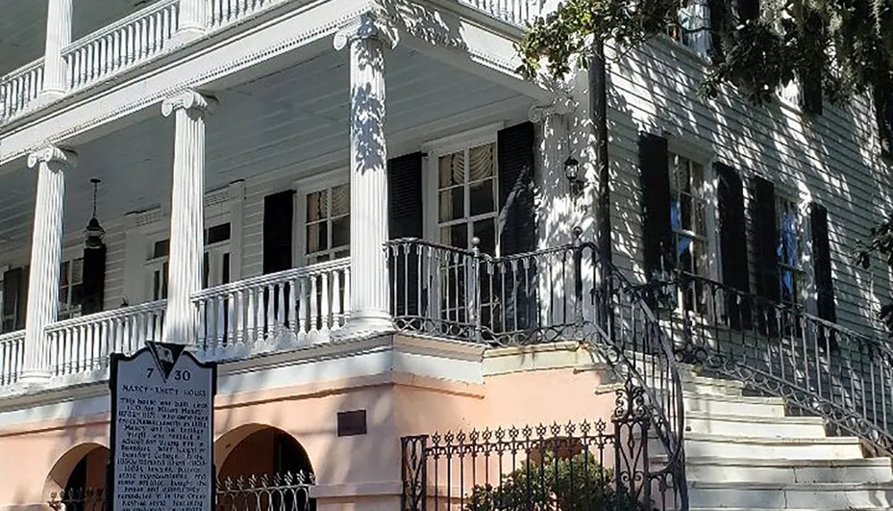This image shows a two-story white house with black shutters front columns external stairs leading to a veranda and a historical marker sign in the front