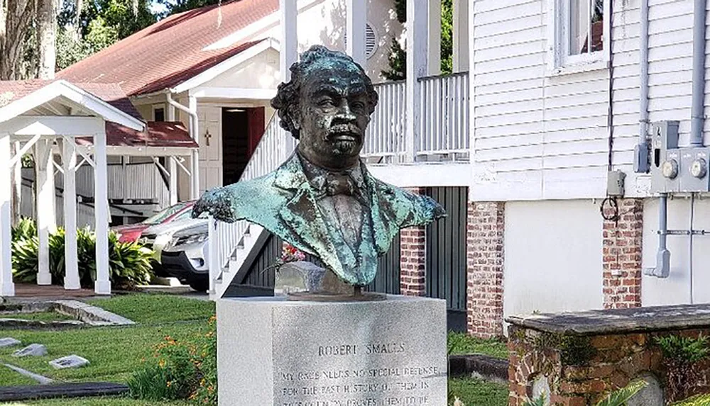 The image shows a bust of Robert Smalls set on a pedestal with an inscription displayed in an outdoor setting near a building with a white porch and a parked car visible in the background