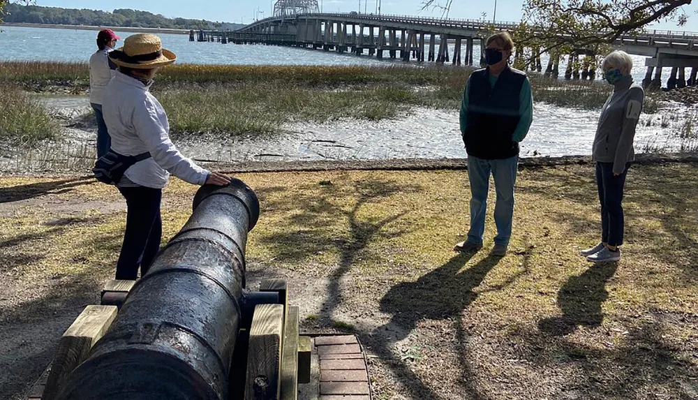 Three people are standing near an old cannon with a scenic water view and a bridge in the background