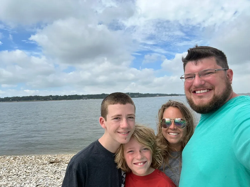 A group of four people are smiling for a selfie with a scenic lake and cloudy sky in the background