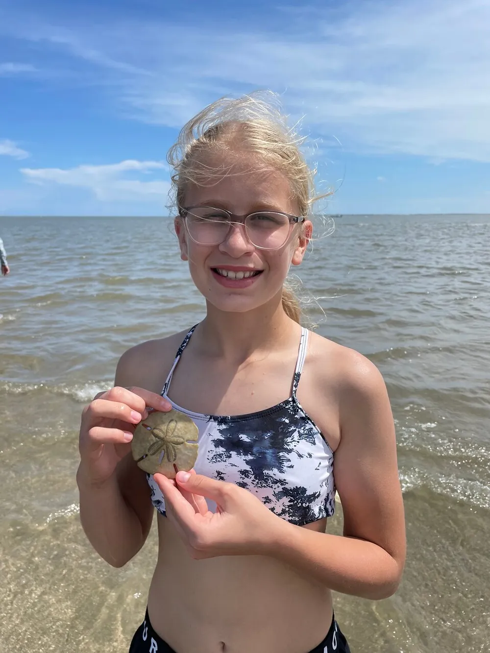 A smiling girl with glasses is holding a sand dollar at the beach on a sunny day