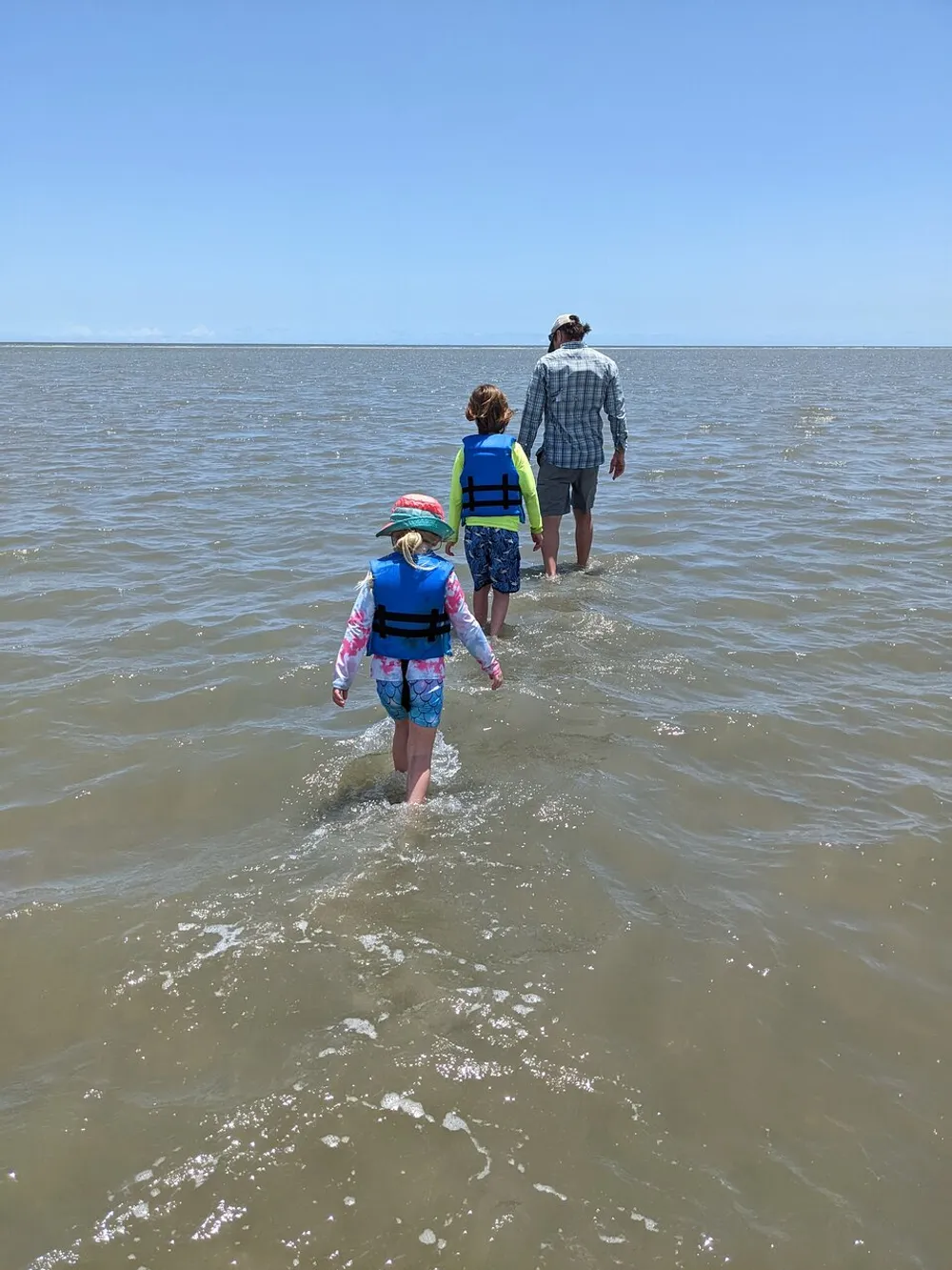 Three individuals possibly a family are walking through shallow waters facing away from the camera on a sunny day