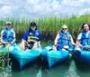 Four individuals are smiling while sitting in kayaks among tall green reeds wearing life jackets under a sunny sky