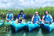 Four individuals are smiling while sitting in kayaks among tall green reeds, wearing life jackets under a sunny sky.