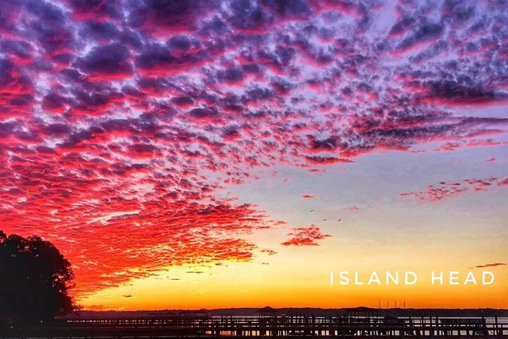 The image shows a vibrant sunset with a dramatic cloud-filled sky casting shades of pink and orange over a silhouetted landscape with a pier and the text ISLAND HEAD overlaid at the bottom
