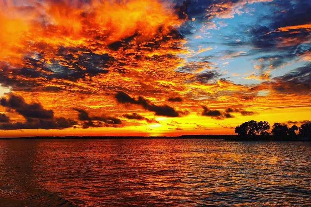 The image shows a vibrant sunset with fiery clouds reflecting over calm waters with silhouettes of trees on an island near the horizon