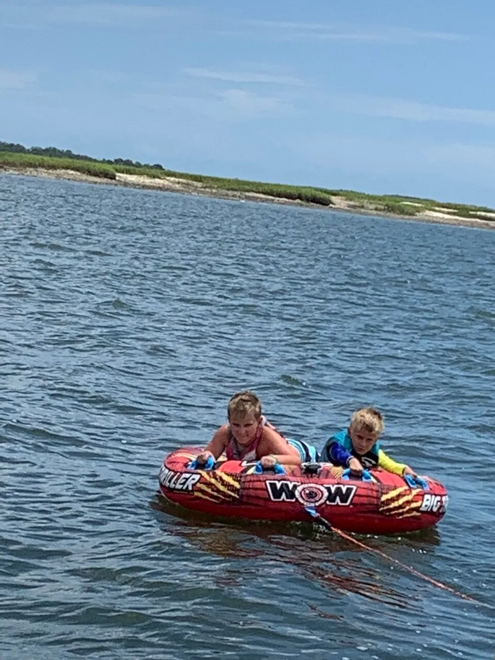 Two children are sitting on an inflatable water tube on a calm body of water with a sandy shore in the background