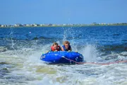 Two people are enjoying a thrilling ride on a water tube being towed across a lake, with splashing water all around them.