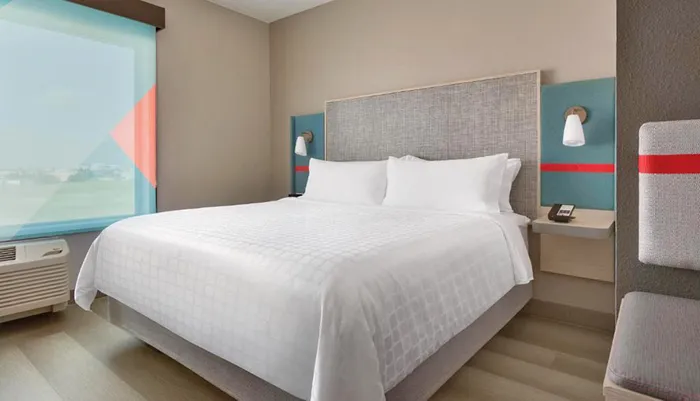The image shows a modern hotel room with a neatly made bed contemporary furnishings and a large window offering natural light