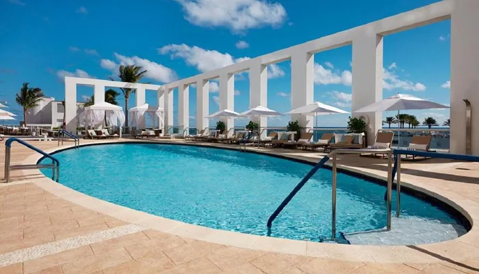 The image shows a serene resort swimming pool flanked by white columns and loungers under a clear blue sky