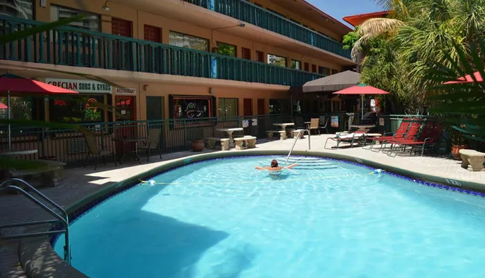A person is enjoying a swim in an outdoor pool surrounded by a motel building with a restaurant nearby on a sunny day