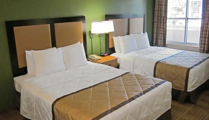 The image shows a tidy hotel room with two double beds a nightstand and a window with curtains