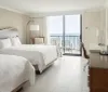 The image shows a tidy and modern hotel room with a large bed minimalist furniture and a balcony overlooking the ocean
