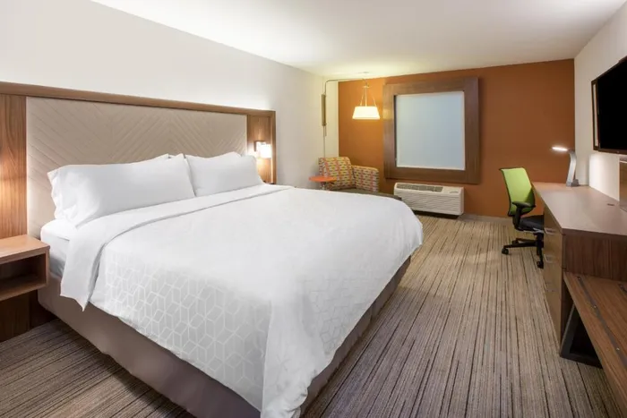 The image shows a neatly arranged modern hotel room with a large bed a desk with a chair a window and a wall-mounted TV