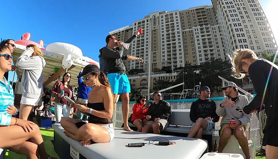 A group of people is socializing on a boat with inflatable toys and drinks, with a large building in the background.