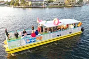 A group of people enjoys a sunny day on a vibrant yellow and white party boat adorned with a large inflatable flamingo on its roof, cruising through a canal lined with houses.