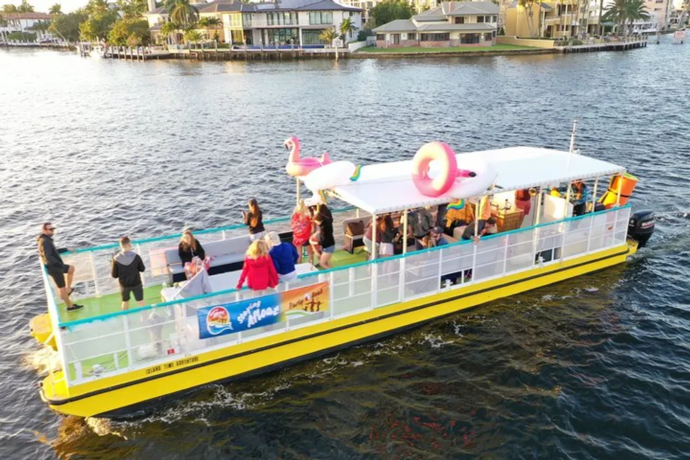 A group of people enjoys a sunny day on a vibrant yellow and white party boat adorned with a large inflatable flamingo on its roof cruising through a canal lined with houses
