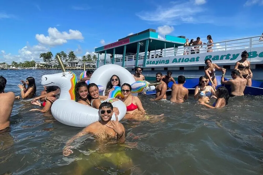 A group of people enjoy a sunny day in the water with various inflatables near a boat labeled Staying Afloat Party Boat
