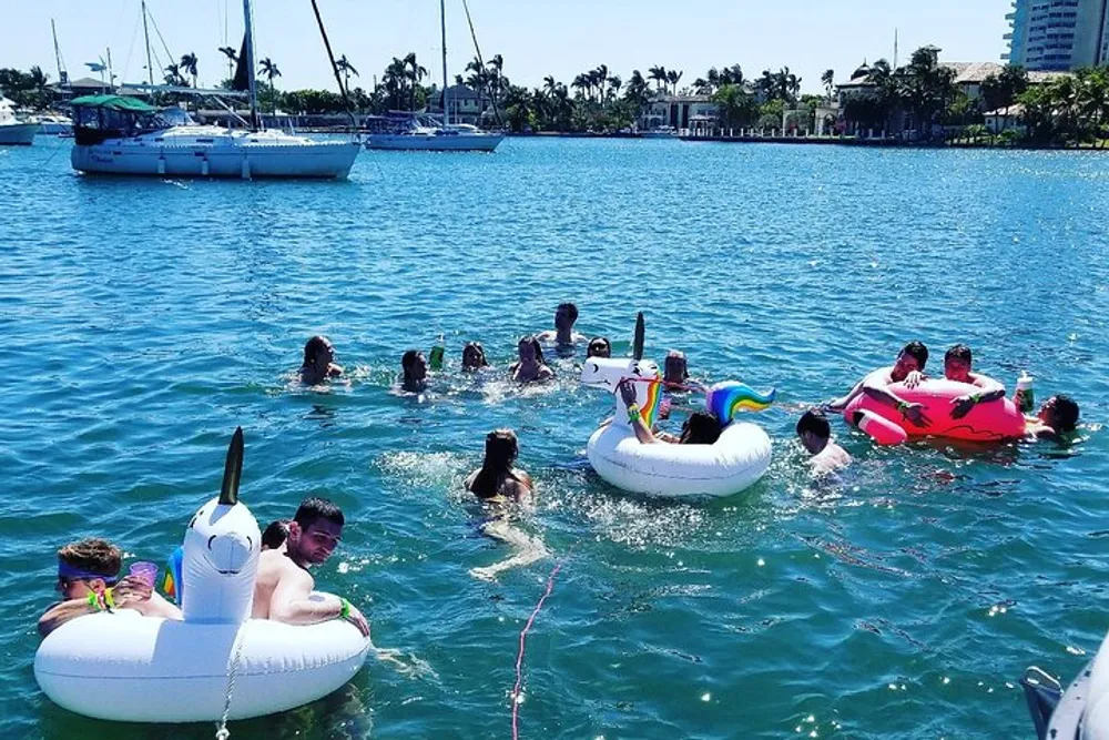 A group of people are enjoying a sunny day in the water with various inflatable swim rings and floats near moored sailboats