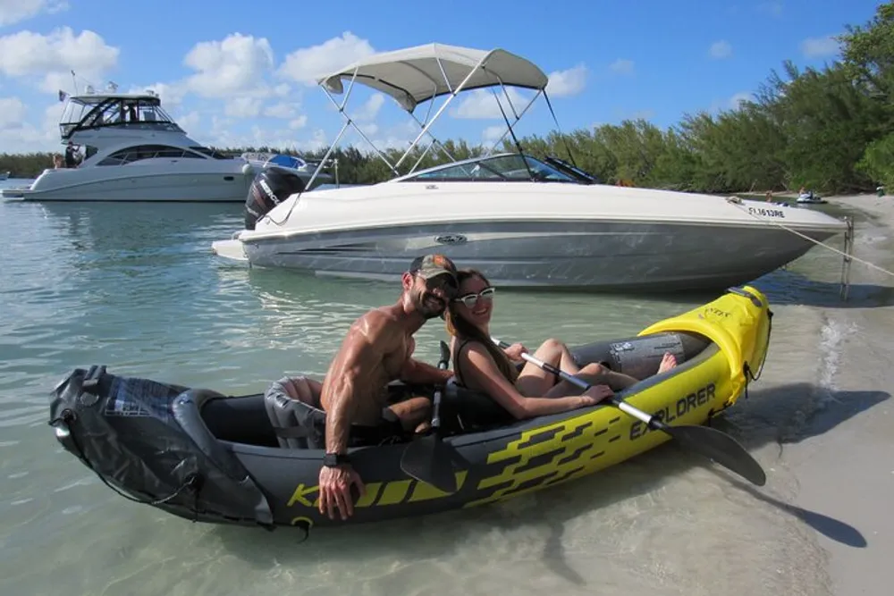 Two people are smiling and sitting together in a small inflatable boat on a clear shallow beach with larger boats visible in the background