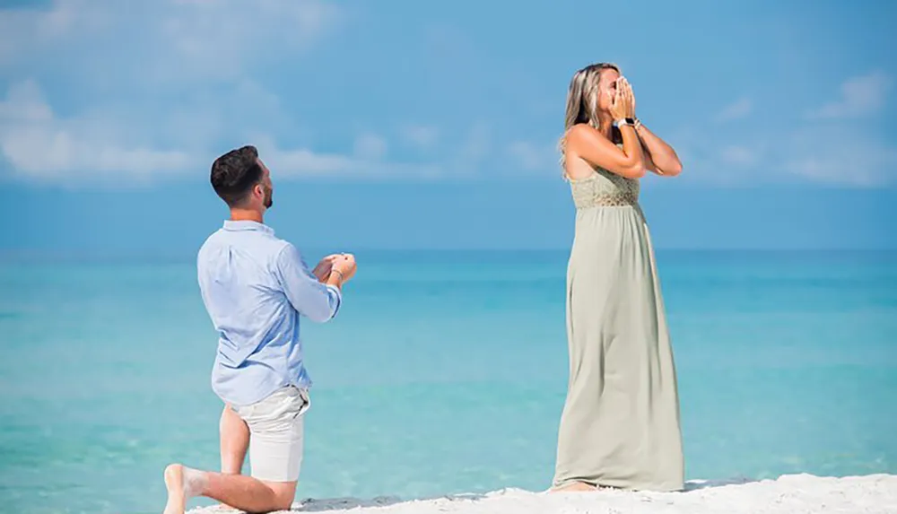 A man is kneeling on a sandy beach holding an engagement ring towards a surprised woman with her hands over her face
