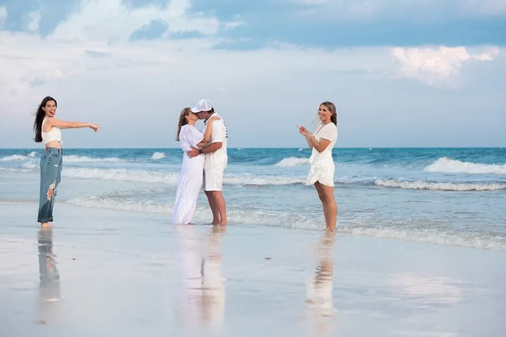 A couple is embracing and kissing on a beach while two women one on either side are smiling and gesturing towards them possibly capturing or celebrating the moment