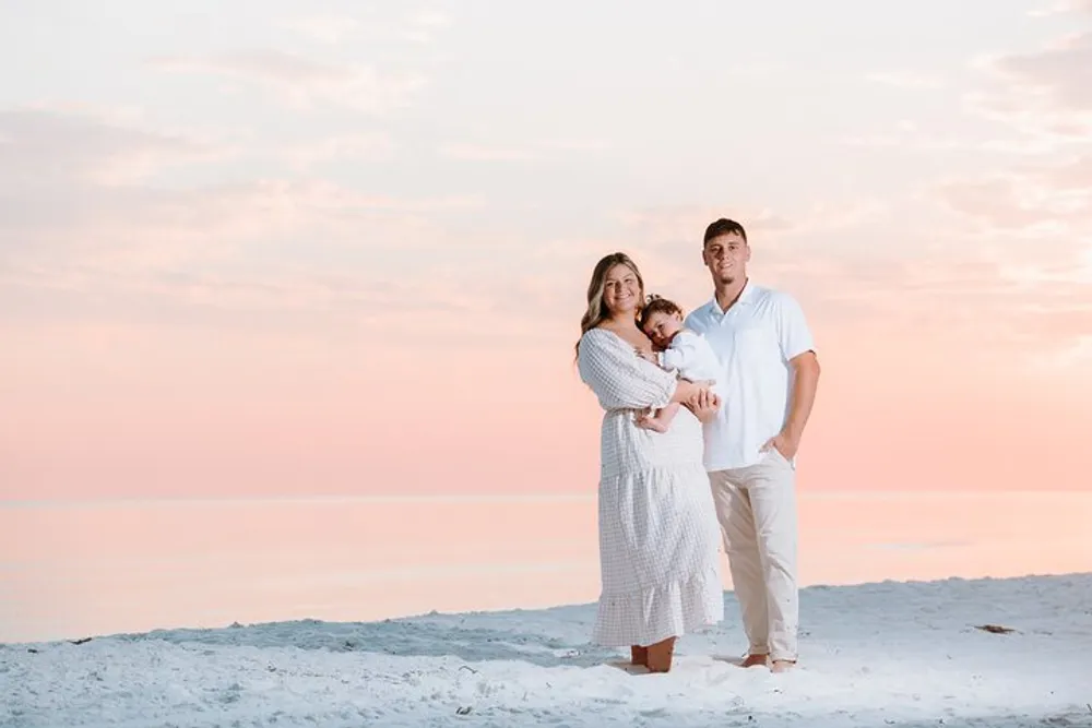 A family with two adults and a small child is standing on a beach posing for a photo against a soft pink-hued sunset