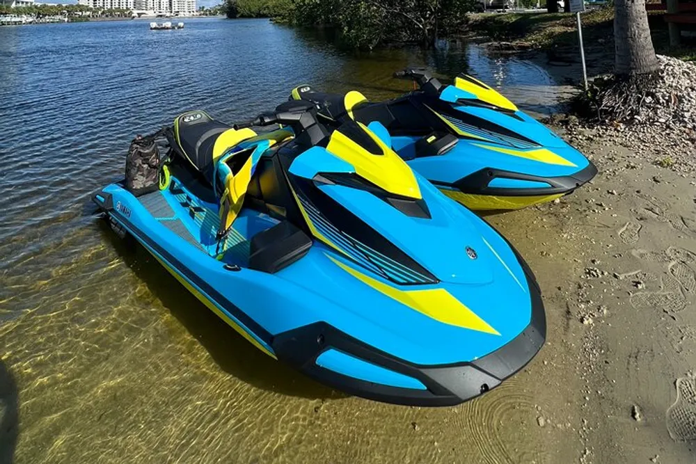 Two blue and yellow personal watercrafts are parked on the shoreline of a clear water body with surrounding greenery and a cloudy blue sky