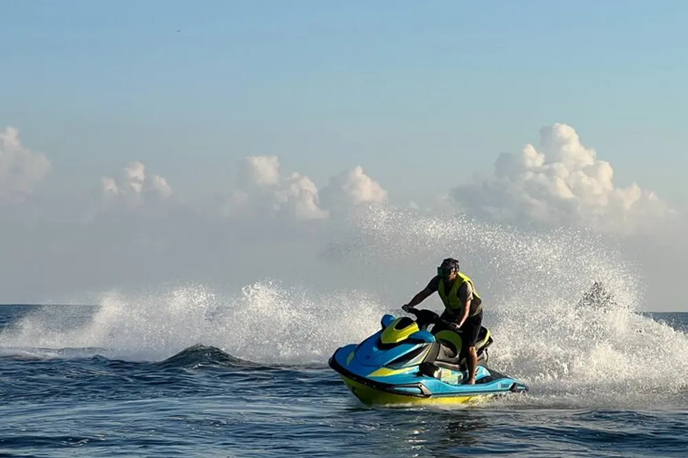 A person is riding a colorful jet ski on the ocean creating splashes against a backdrop of blue sky and white clouds