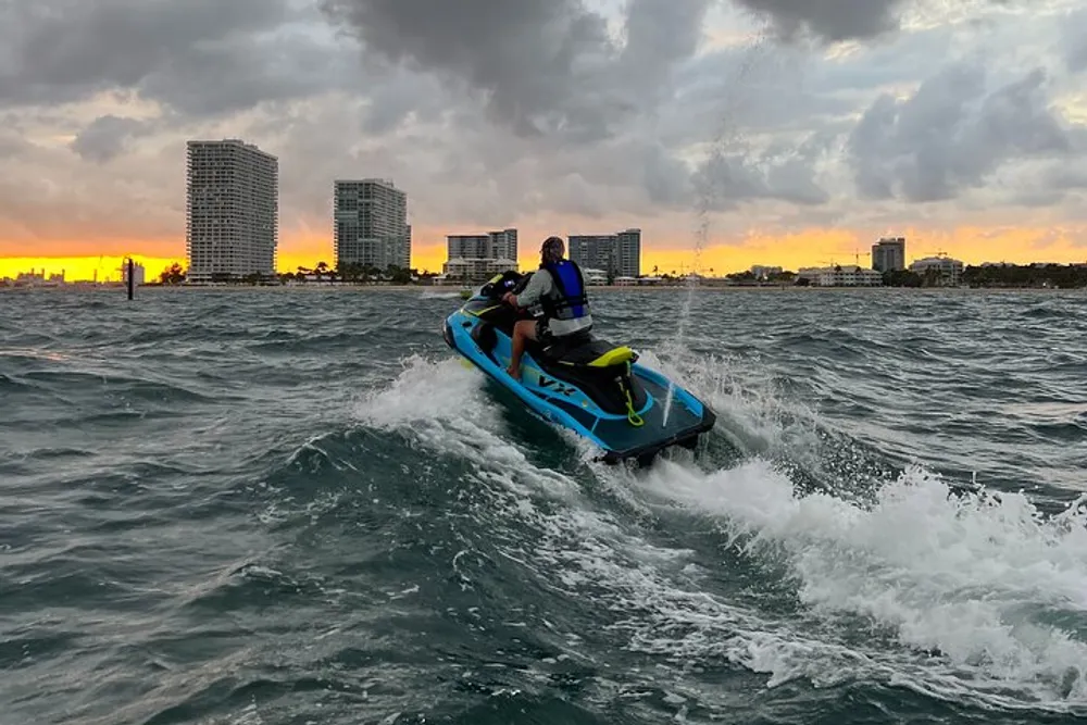 Two individuals are riding a jet ski on choppy waters with a vivid sunset and city skyline in the background