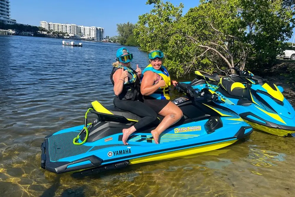 Two people are smiling and posing on a blue and yellow jet ski near the shore with trees and buildings in the background