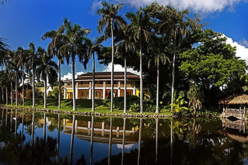 The image depicts a stately two-story building with a large portico surrounded by tall palm trees and reflected in a tranquil body of water in the foreground