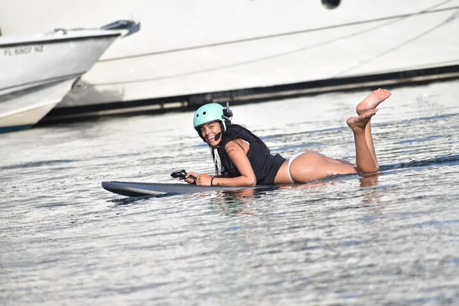 A person is lying on a wakeboard in the water, smiling and holding a handle attached to a rope, wearing a helmet, likely after or during a playful fall.